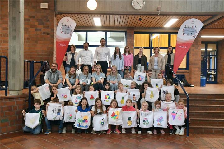  Gruppenbild mit Landrat Kai Zwicker (stehend, Zweiter von links) sowie DRK-Vorstand Dr. Cornelius Rack (daneben), Betreuungspersonal in der Aula der Jodocus-Nünning-Gesamtschule, und die Kinder zeigen stolz ihre bemalten Jutetaschen.